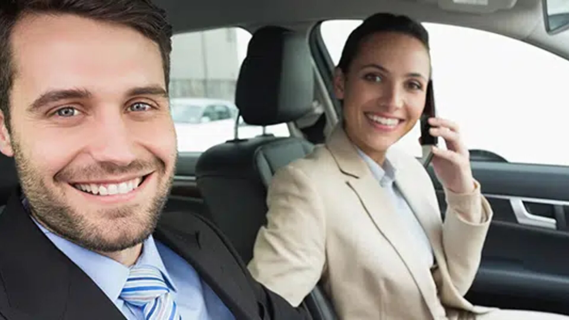 Smiling businessman and businesswoman in a car, woman on phone, man looking at the camera