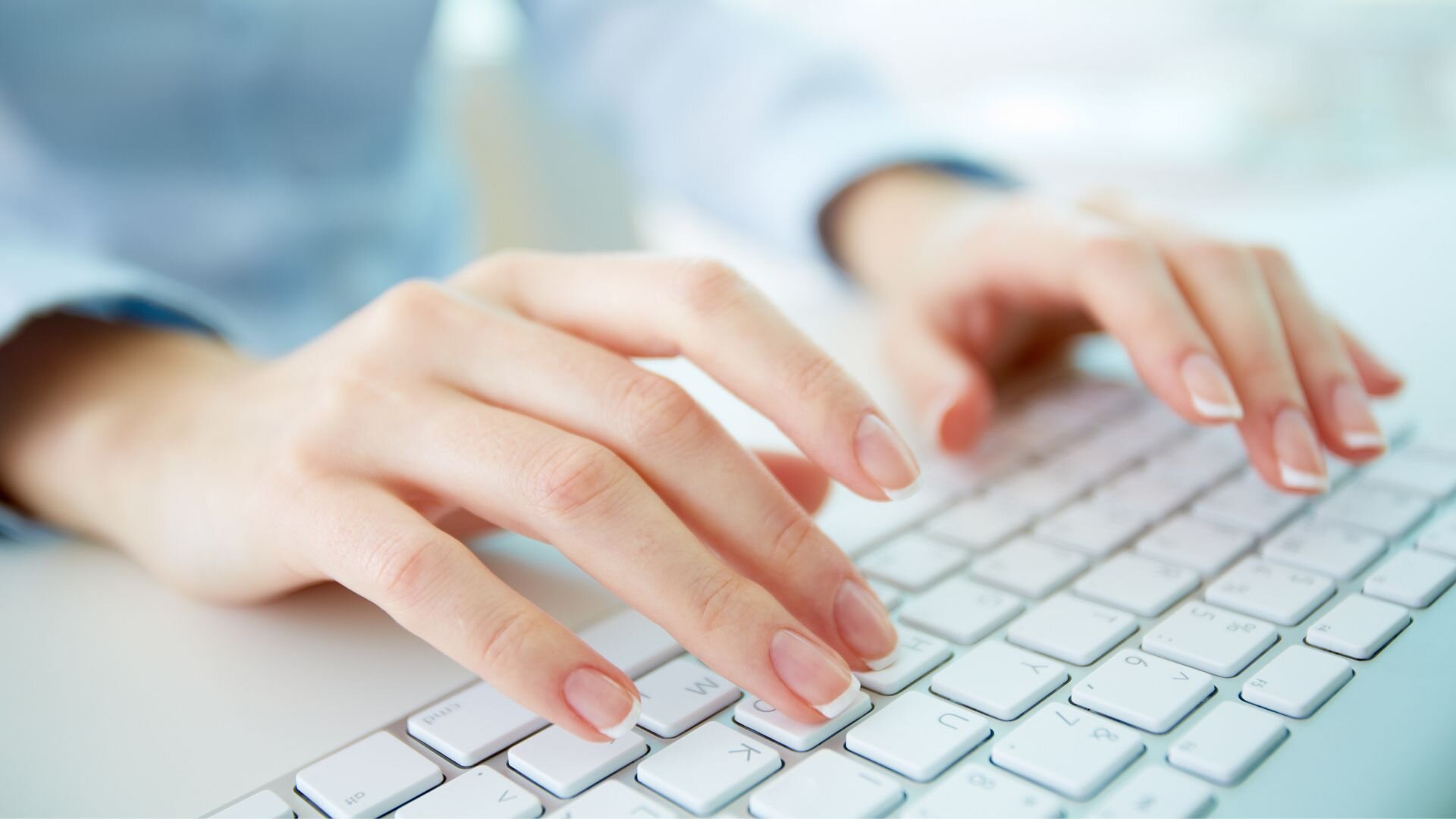 a woman's hands typing on a white computer keyboard, with a bright, softly blurred background